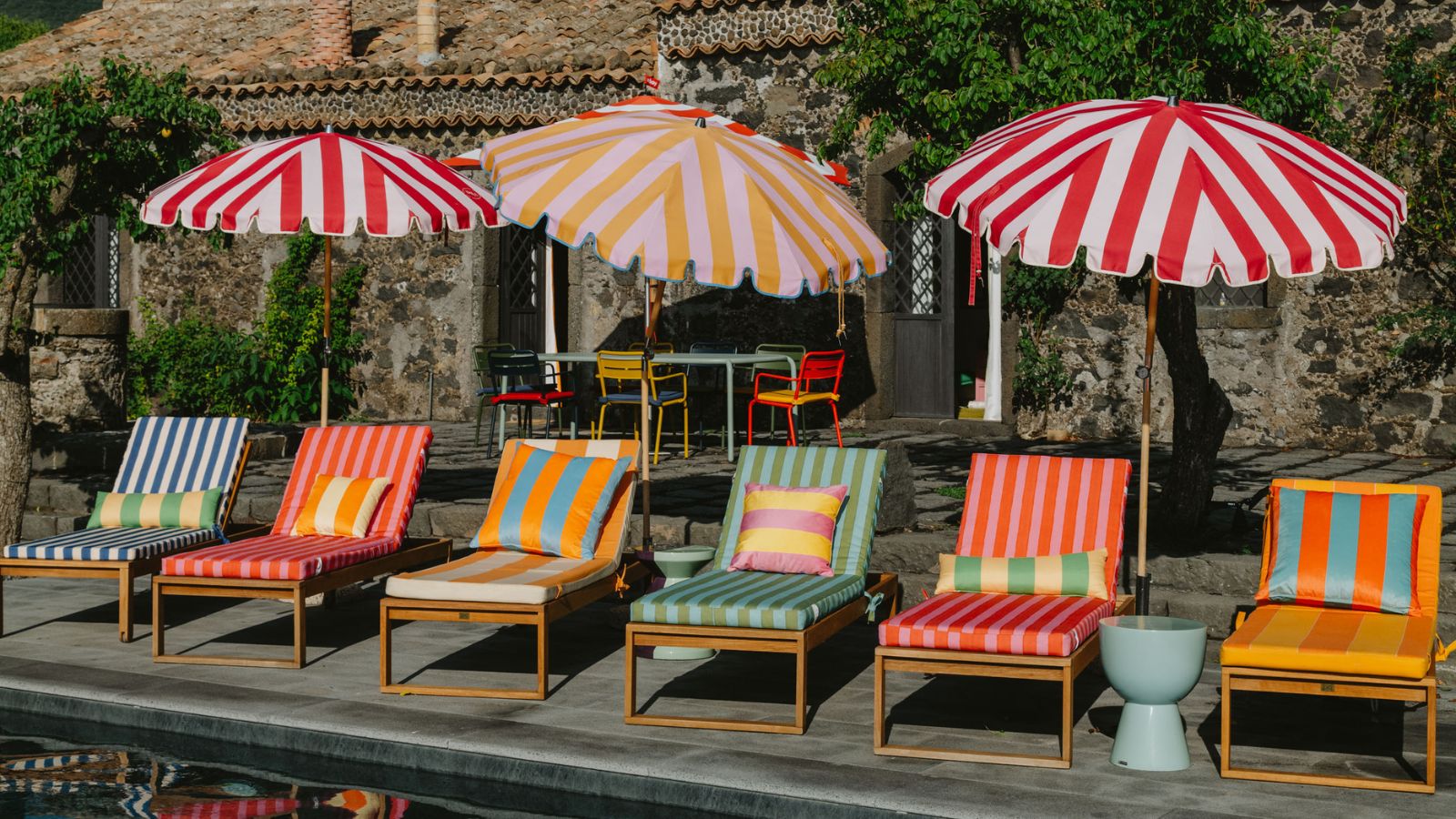 colorful striped pool loungers with striped patio umbrellas by a pool with a stone building and trees in the backdrop