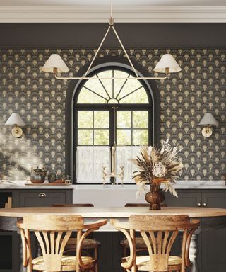 A black kitchen with art deco wallpaper, a white hanging light with two lampshades, black cabinets and a gold faucet above a white sink, and a curved light wooden dining table with flowers on and two chairs