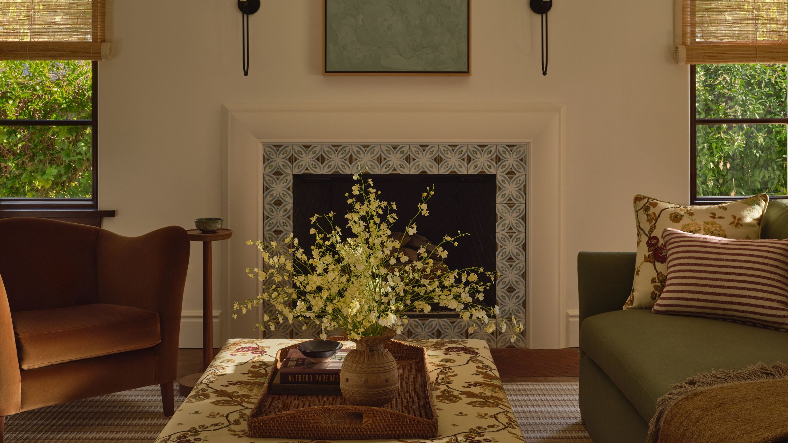 a neutral living room with Spanish style tiled fireplace, two large windows and floral upholstered ottoman