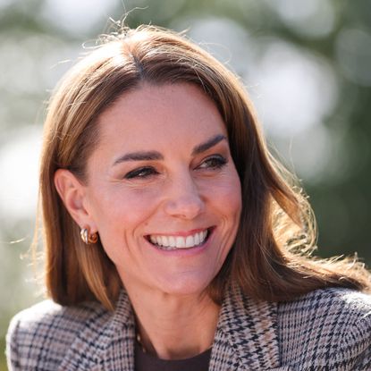 Princess Kate wearing a plaid blazer and smiling in front of a tree