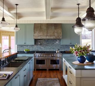 Classic kitchen with coffered ceiling, multi ceiling pendants hung over island and sink, and sage green cabinets and tiled backsplash