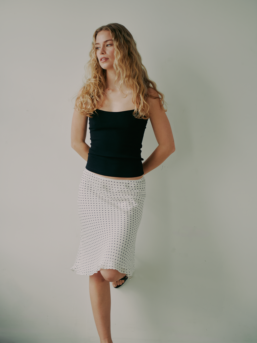 Elle Chapman stands with one foot against the white wall behind her. She is wearing a black tank top and white, knee-length skirt with black polka dots.