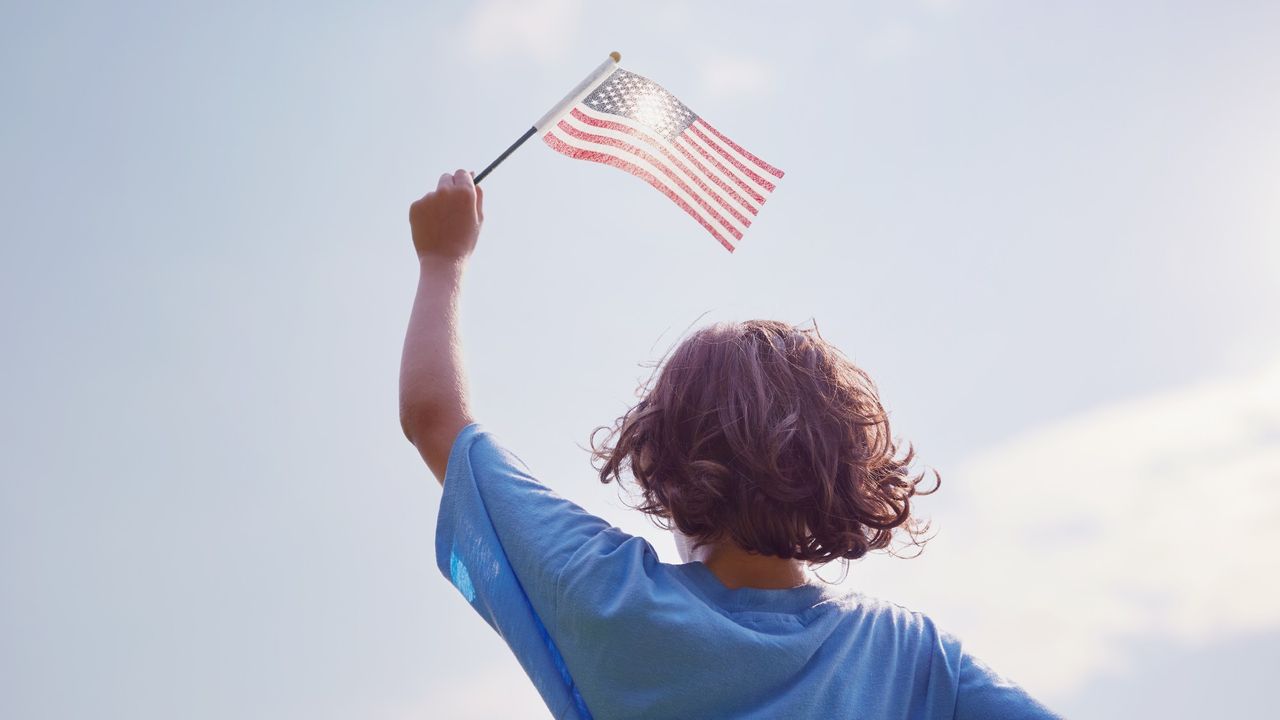 Silhouette of a little boy from the back holding an American flag against the sky