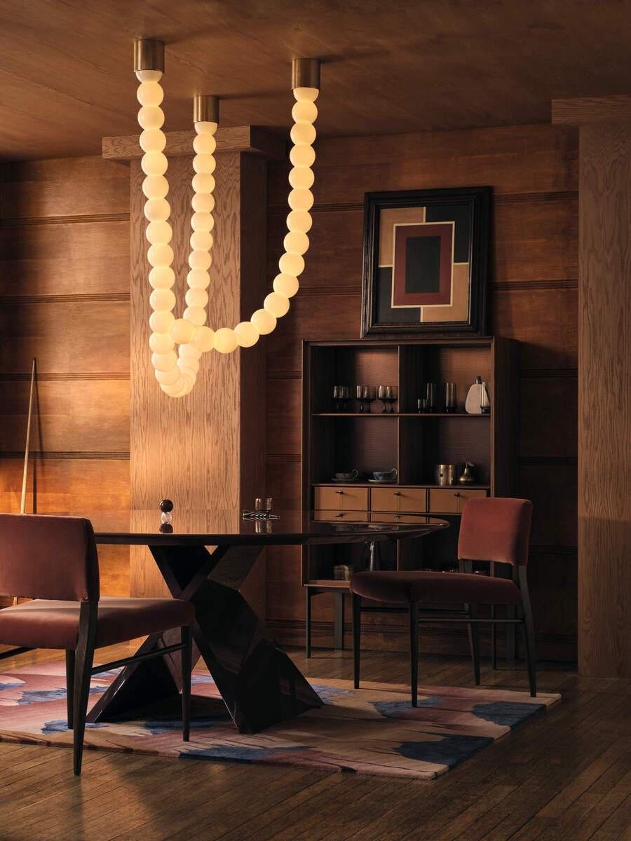 Wood-drenched dining room (the walls, floor, and ceiling are all a rich warm wood). There is a geometric rug on the oval dining table. Above the dining table hangs a statement pendant light that resembles a beaded necklace.