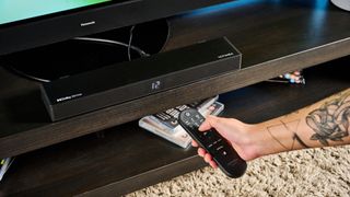 the ultimea nova s50 a black compact soundbar with LED on the front and a wired subwoofer is photographed on a black TV mount connected to a panasonic TV