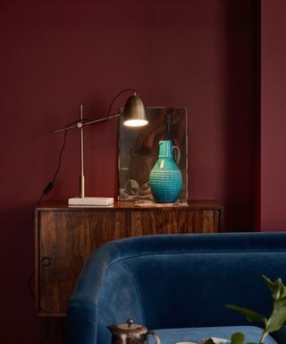 A small brass leaver lamp on a dark wood console unit in front of a deep, berry red wall. Stood beside a small blue ceramic jug and a dark abstract painting.