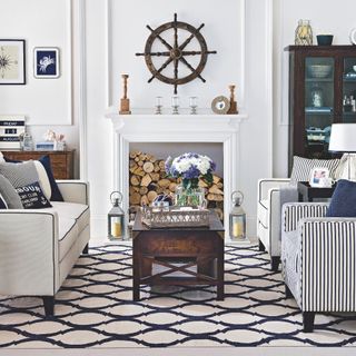 white and blue nautical living room with vintage pieces, white and blue sofa, blue striped chairs, wooden coffee table, ship wheel, logs in fire, artwork, storage, lanterns