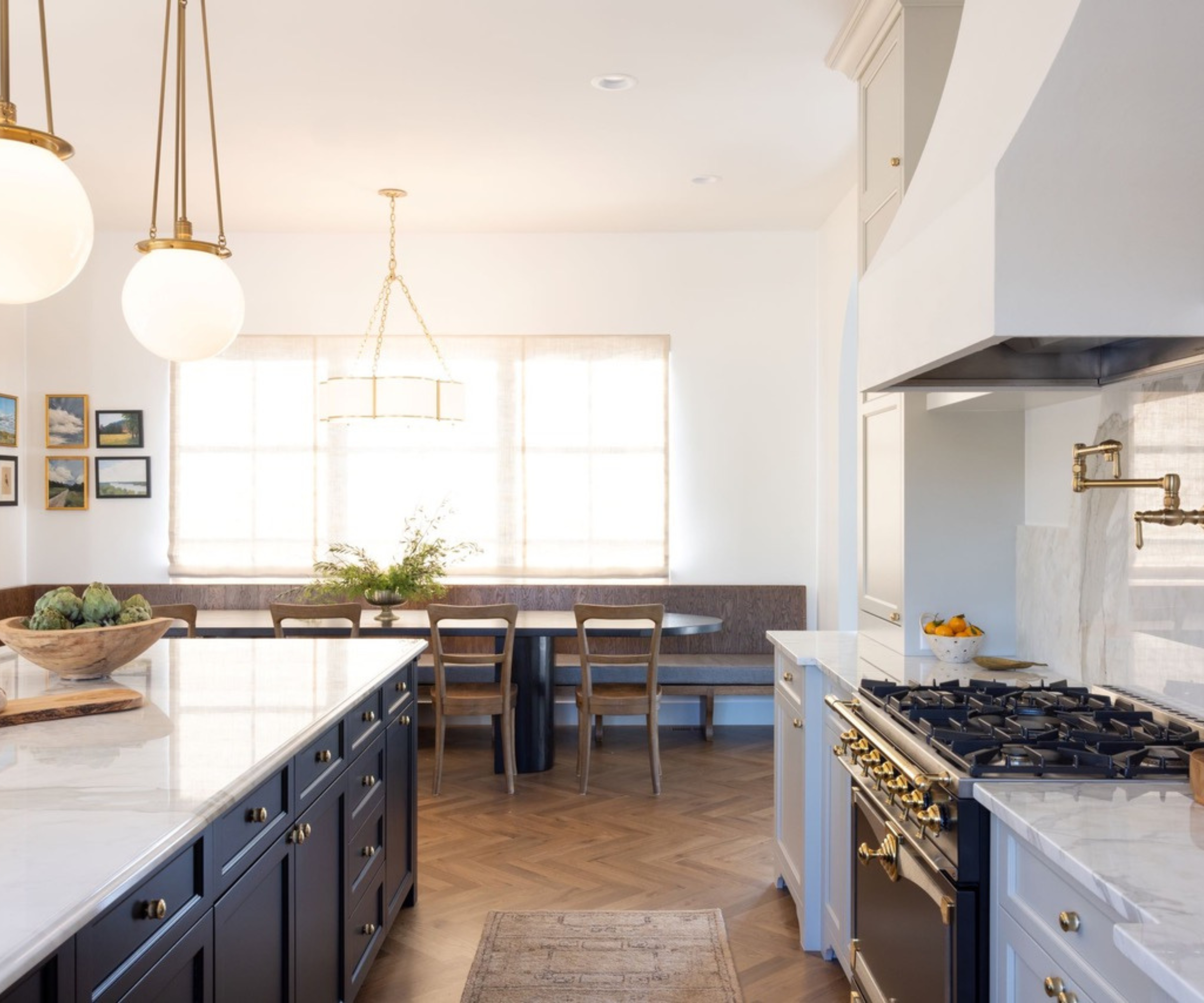 An expansive, airy kitchen featuring navy blue island cabinetry with white marble countertops, herringbone wood floors, and a breakfast nook with a built-in banquette under large windows