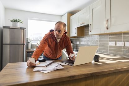 A wide angle shot of a man standing and leaning against his kitchen worktop at home. He is using his laptop to process online banking and asses his personal finances. He looks stressed out.
