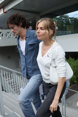 Sienna Miller walking wearing a white V-neck sweatshirt, black jeans, white mini bag and her blonde hair in a claw clip