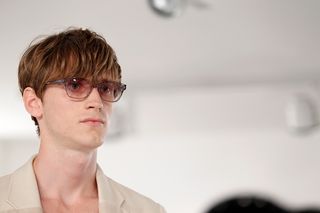 male model to the left of the frame wearing a beige blazer, long fringe and glasses