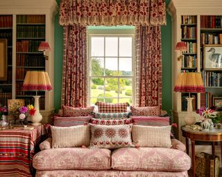 Traditional, colorful living room full of pattern and texture, large floor to ceiling window dressed with floral drapes with valance, blue painted walls, two matching white bookcases, pink floral patterned sofa, wooden side tables either side with table lamps with decorative shades