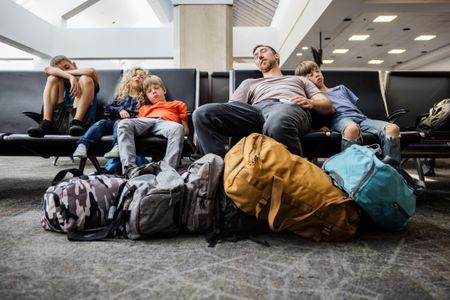 A family with multiple children traveling together at an airport, waiting to board their plane. They sleep in a waiting area, surrounded by their luggage, bags, and carry-on items.