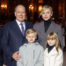 Prince Albert, Princess Charlene, Prince Jacques and Princess Gabriella wearing coats and scarves standing outside a lit-up building