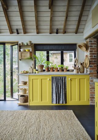 country grandmillennial kitchen with yellow cabinets, curtain, baskets, natural rug, beams, plants pots