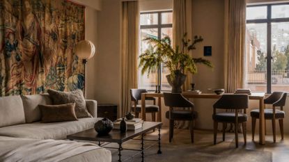 A living room with a tropical tapestry on the wall behind a gray couch near a floor lamp and a dining set with a vase of feathered stems