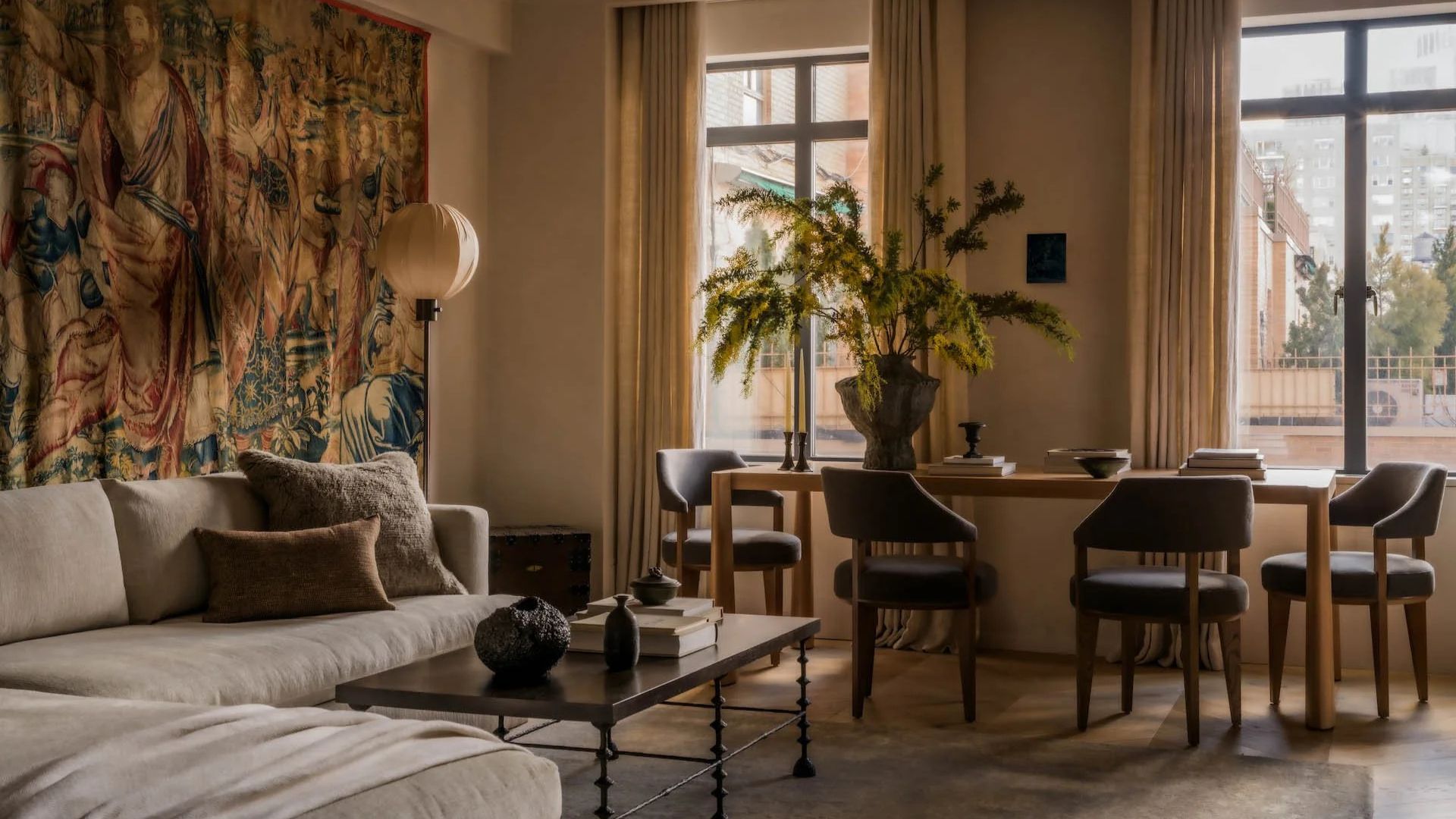 A living room with a tropical tapestry on the wall behind a gray couch near a floor lamp and a dining set with a vase of feathered stems