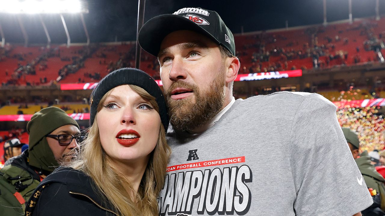 Taylor Swift wearing a black beanie and hoodie hugging Travis Kelce, who is wearing an AFC Champions shirt and black hat on the field after the 2025 AFC championship game