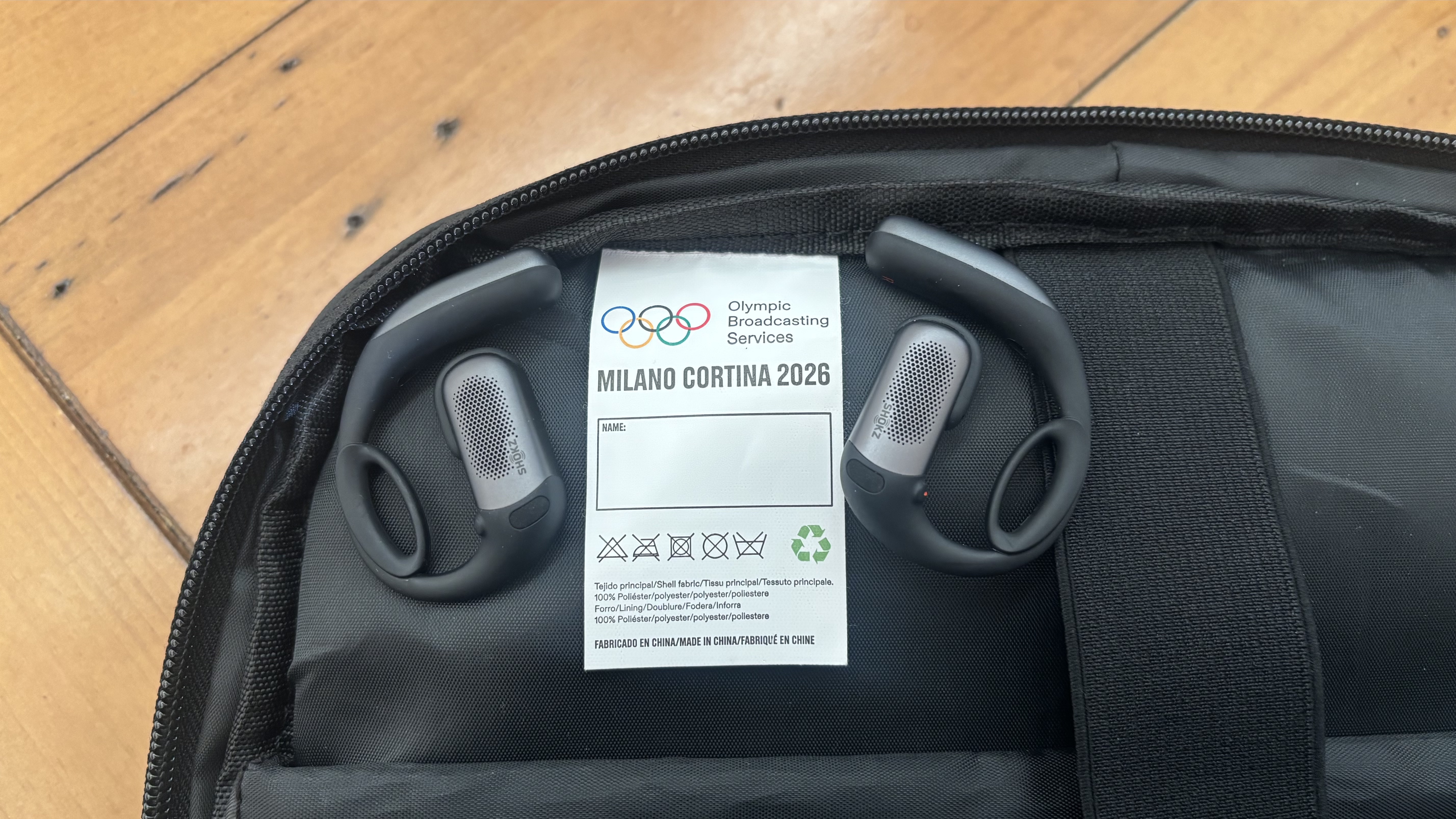 Shokz OpenFit Pro open earbuds placed on the inside of a black rucksack, featuring Milano Cortina 2026 Olympic Games logo.