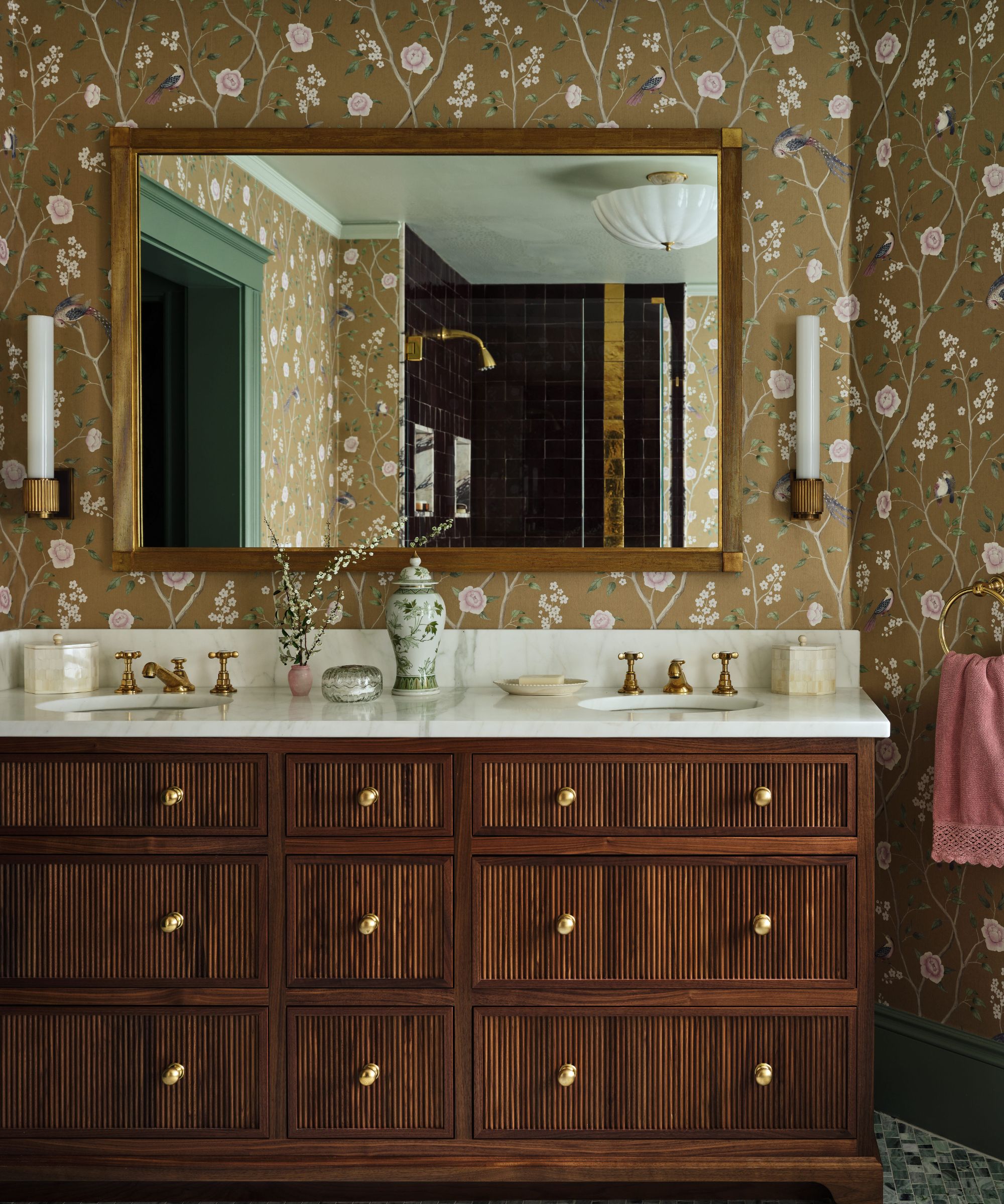 Double-sink bathroom vanity with dark wood fluted cabinets, white marble top, and a large gold-framed mirror against brown floral wallpaper