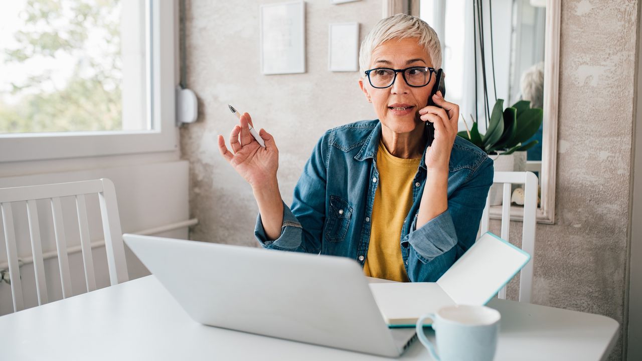 An older woman talks on the phone with her laptop open at the kitchen table.