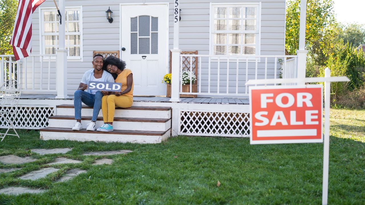 Young couple sits in front of a house they have purchased, holding a sign that says &quot;sold&quot;