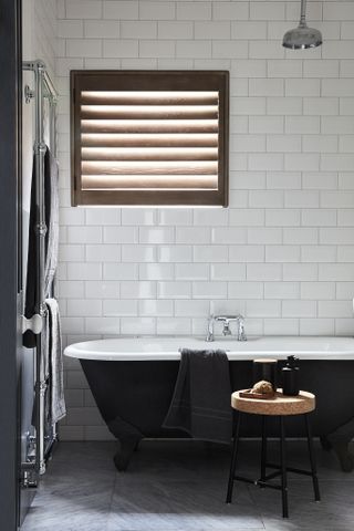 white bathroom with black roll top tub, white metro tiles, grey stone flooring, towel rail, shower over tub, shutter on window