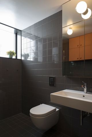 Interior view of a bathroom at the Oslo family house featuring dark grey tiled walls and flooring, a window with a plant, a white toilet, a white sink and a wall mounted mirrored cabinet
