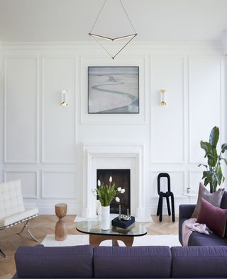 A white living room with molding on the walls and wooden floors. There is a fire place in the middle of the back wall with a black accent chair beside it. There is a large, indigo sectional sofa on top of a white rug with a wood and glass coffee table in front of the sofa.