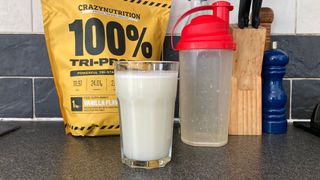 Glass of white protein shake next to protein shaker and bag of Crazy Nutrition Tri-Protein