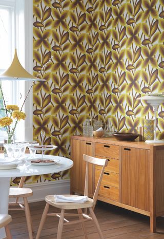 Retro style floral wallpaper above 70s style sideboard, cropping through to white round dining table with wood chairs.
