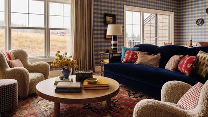 an open plan living area in a home in the Catskills with blue gingham wallpaper, a navy velvet couch, vintage red rug, sheepskin armchairs and a round wooden coffee table