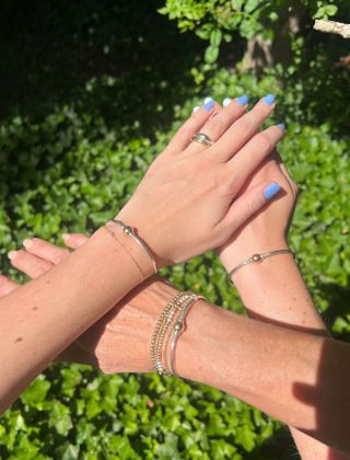 Three hands wearing silver-and-gold Cape Cod screwball bracelets in front of a green bush.