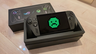 A black ROG gaming handheld console sits in an open black box on a wooden surface. The box displays controller graphics with vibrant icons.