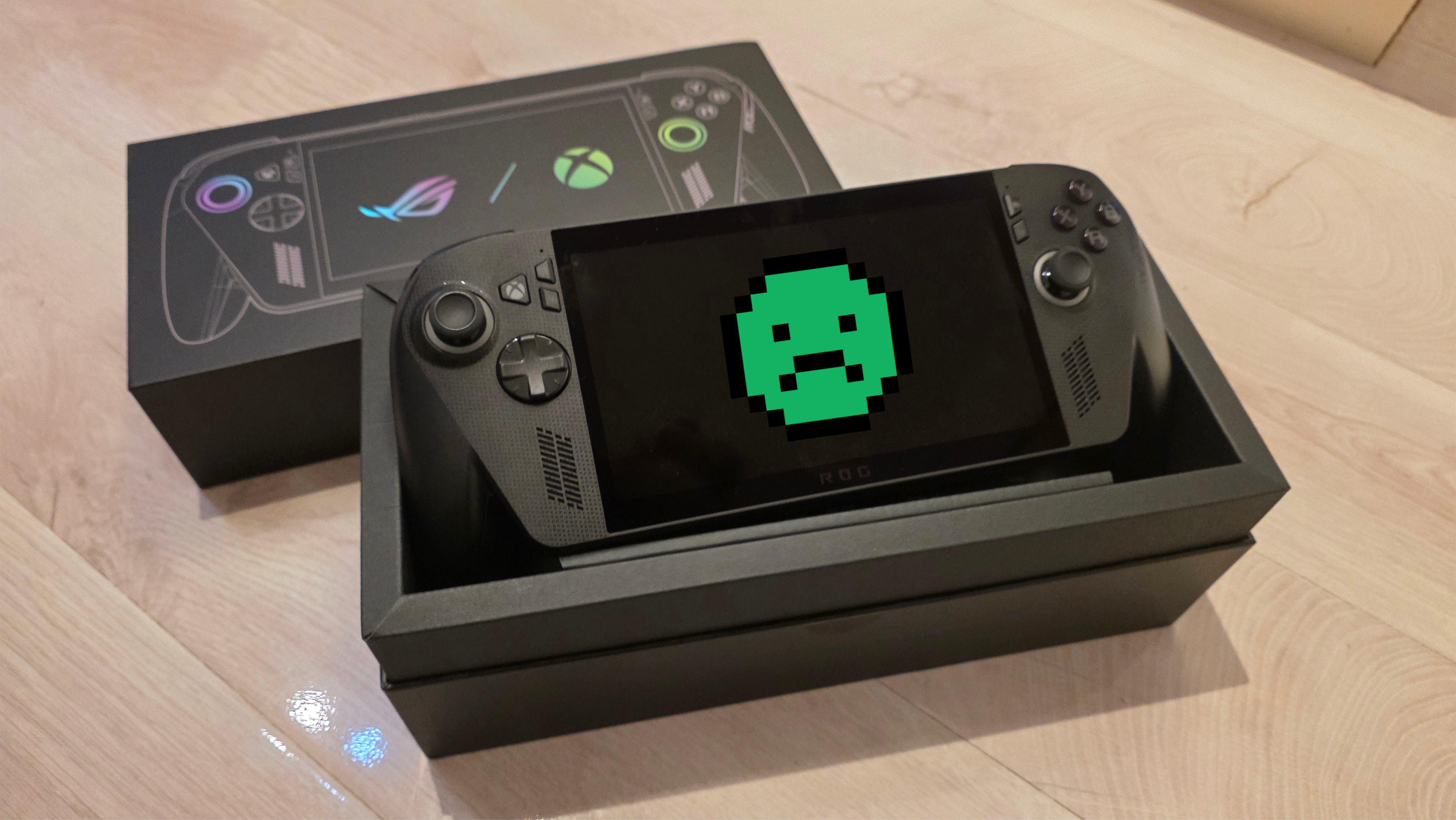 A black ROG gaming handheld console sits in an open black box on a wooden surface. The box displays controller graphics with vibrant icons.