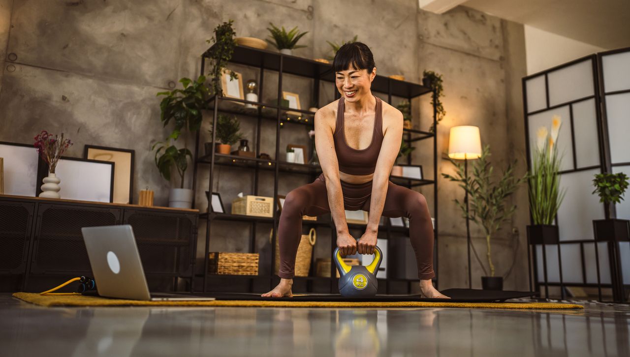 Woman exercises using kettlebell at home. She is barefoot, wearing matching brown crop top and leggings, and she is looking at a laptop on the floor. 