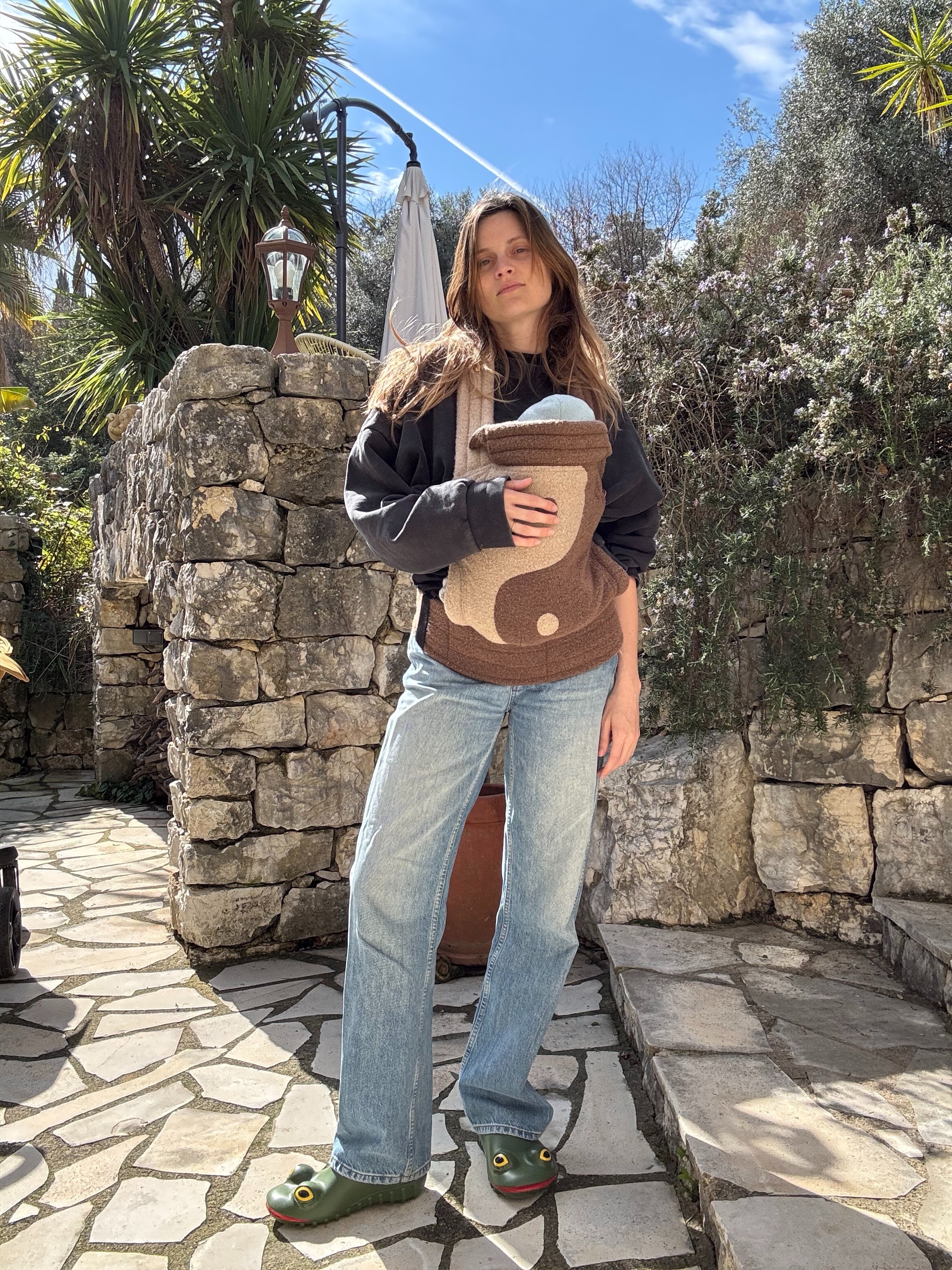 French editor and write Eugenie Trochu poses with a yin and yang baby carriers, a sweatshirt, straight-leg jeans, and JW Anderson frog rain clogs