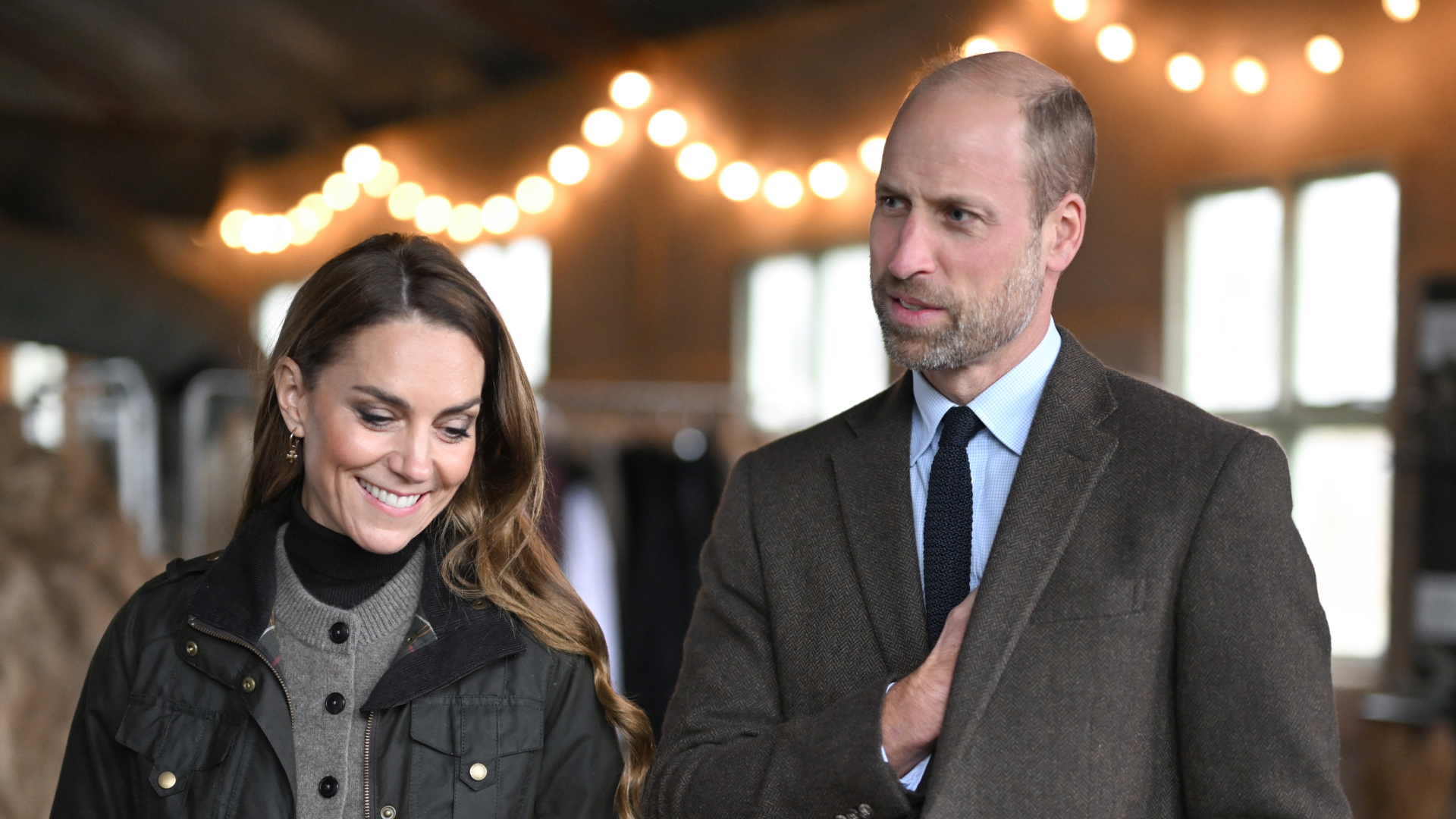 Prince William and Princess Kate wearing brown outfits standing in front of white lights