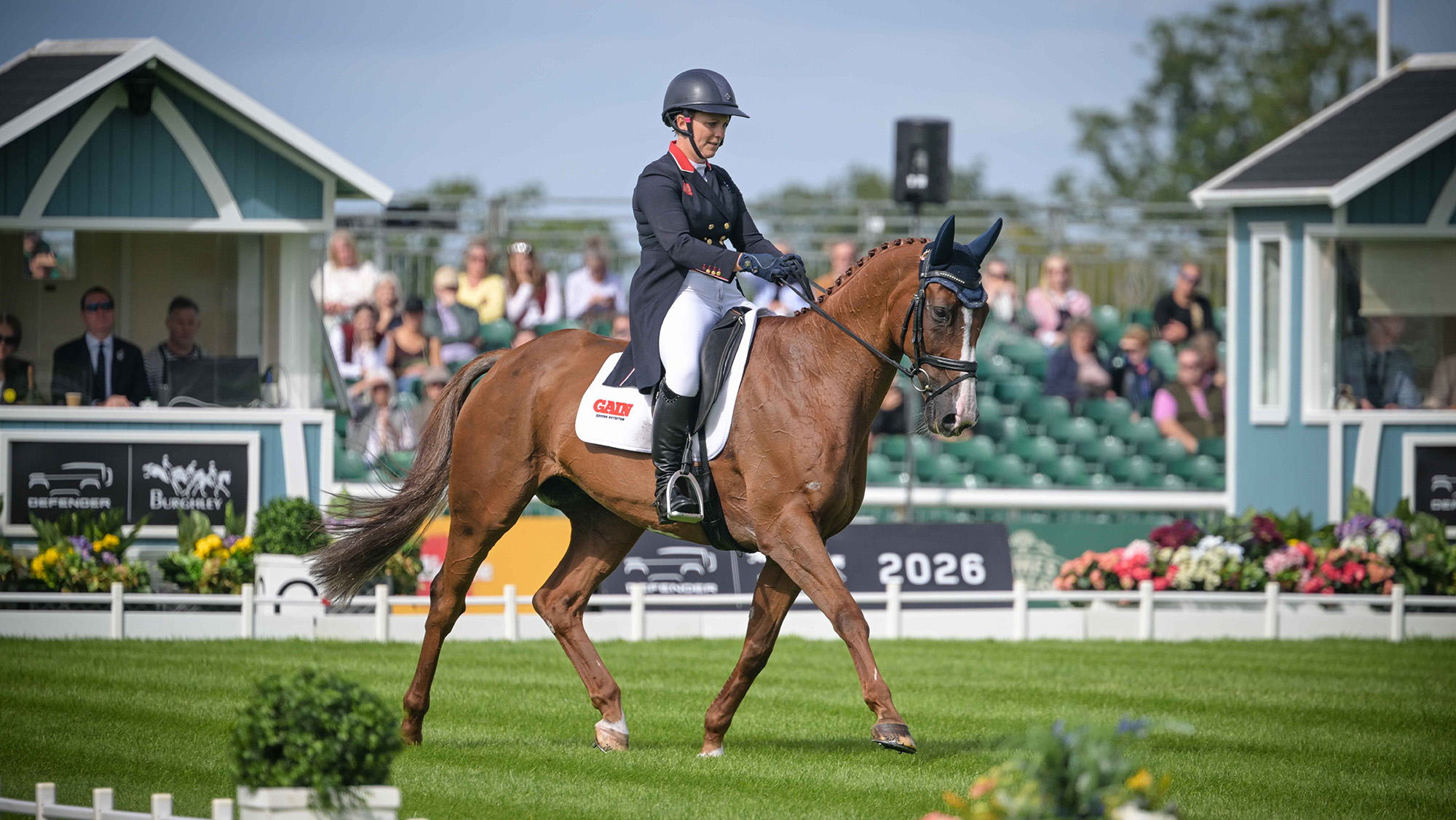 Gemma Stevens, a rider in a dark tailcoat with a red collar, rides chestnut horse Chilli Knight in the Burghley dressage arena