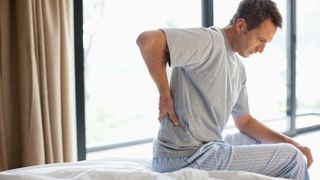 A man sat on the edge of his memory foam bed after waking up with back pain, holding his back