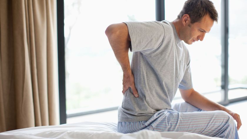 A man in a grey t shirt sits on the edge of his mattress while clutching his lower back after waking up with back and shoulder pain