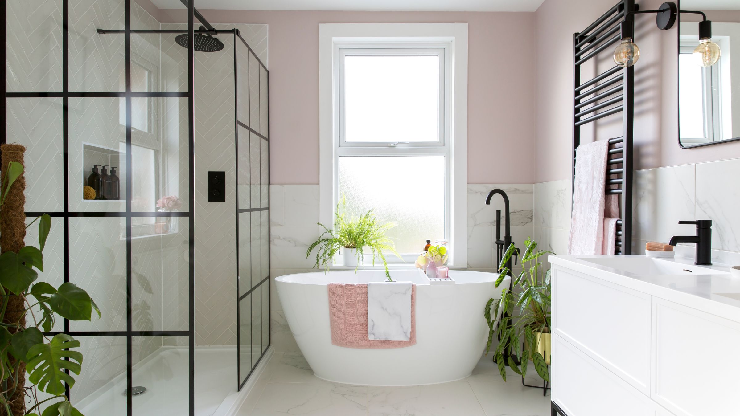 Pink painted bathroom with white marble tiles on the lower part of the wall, a freestanding bath in the middle of the room, and a sink and shower either side