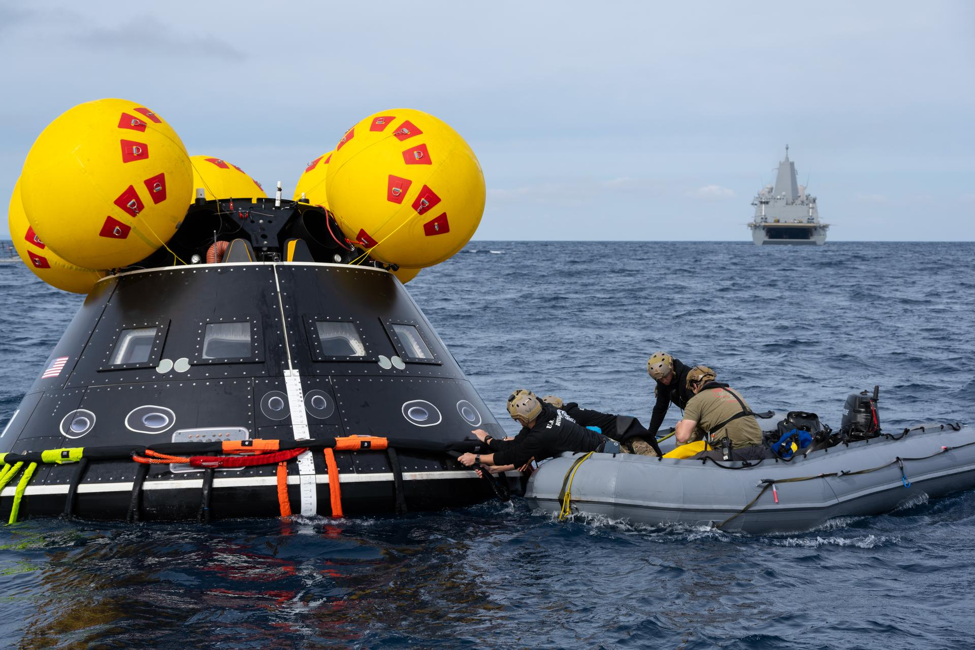 a black space capsule floats in the ocean. Its top is crowned with several inflated yellow balls. Four individuals in a grey inflatable boat are facing the capsule. A couple are securing something with their hands on the capsule. In the distance, a grey NAVY ship floats on the horizon.