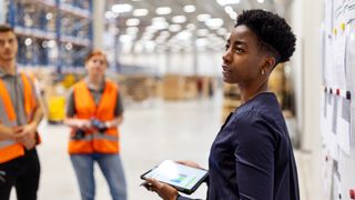 A supervisor stood in a warehouse speaking to workers in high-vis vests, while holding a tablet displaying stats about the supply chain, to represent supply chain oversight.