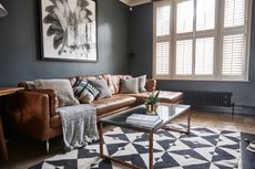 Jason Traves house: Living room with dark grey walls, tan leather sofa, minimalist coffee table with glass top, copper pendant light and black and white pattern rug over a wooden floor