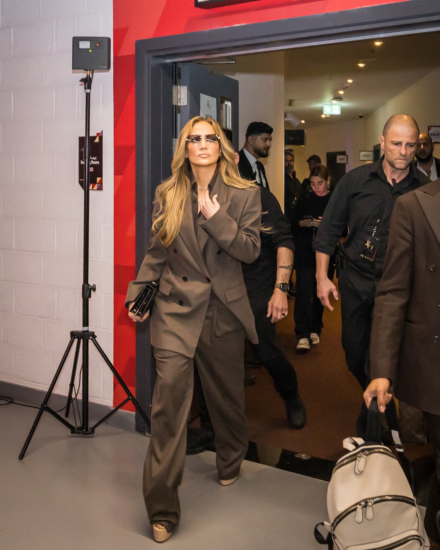 Jennifer lopez wears a brown blazer, brown button-up shirt, black clutch, brown pants, and platform heels.