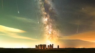 The ancient monument Stonehenge is pictured at night with the Milky Way glowing in a band tumbling towards the horizon. Pereid meteor trails can be seen arcing towards the historic site in a night sky dotted with stars.