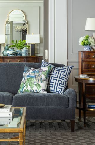 living room with gray/blue sofa, gray textured rug, mahogany antique furniture, gold coffee table, printed green and blue cushions, gray walls, view to entranceway, mirror, console table