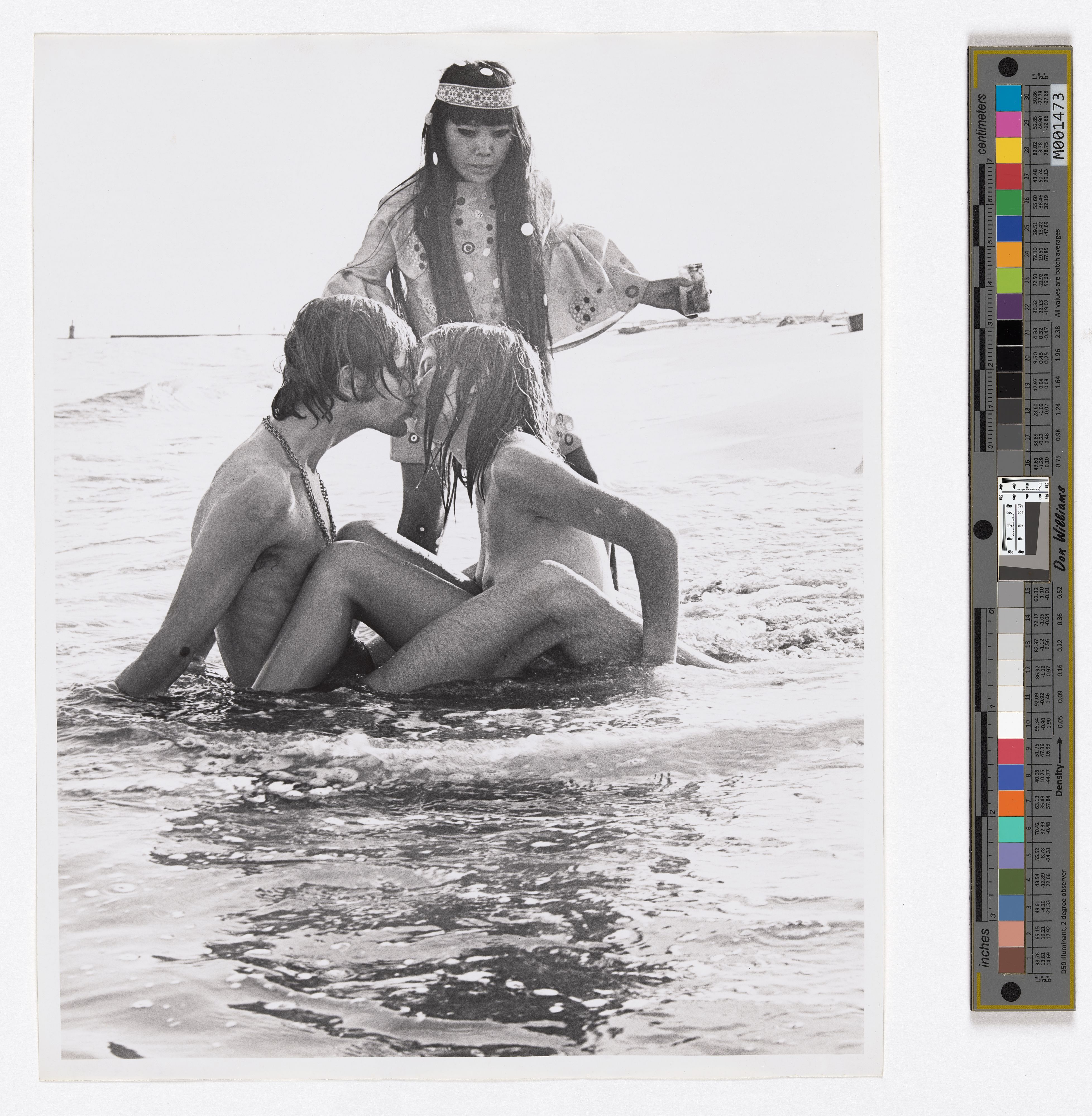 A naked man and woman kissing in the shallows of the sea while a woman in a kaftan-like dress and headband looks on