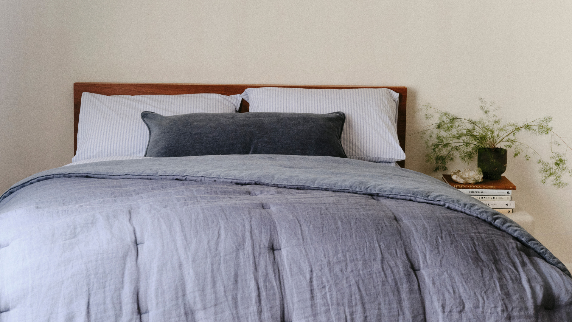 A bed dressed in light blue bedding beside a bedside table with a potted textured plant on a stack of books